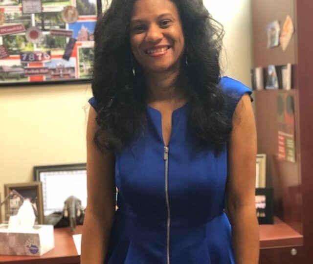 Photo of Kinyata Brown, a black woman with long hair wearing a blue dress. She is standing in her office